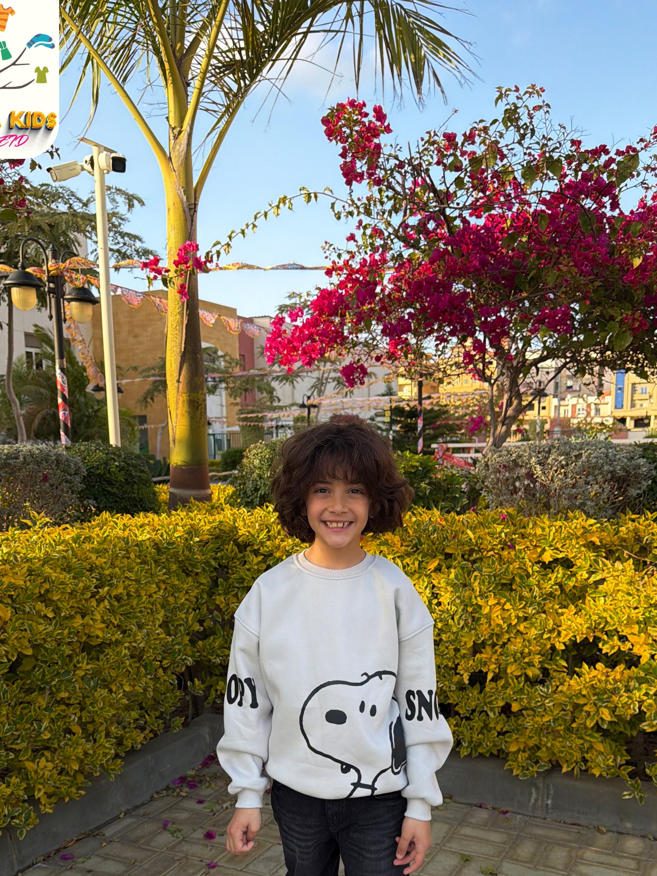 Child wearing a Snoopy sweatshirt standing in front of a garden with yellow bushes and pink flowers.