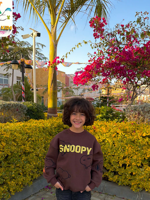 Child wearing a Snoopy sweatshirt standing in front of flowering bushes and palm trees.