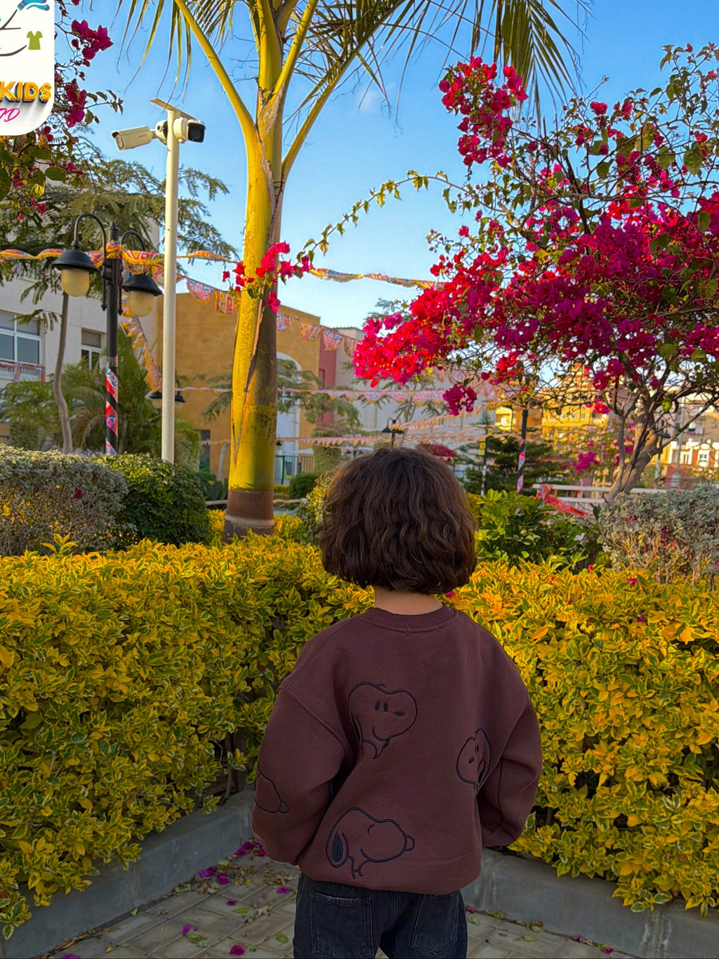 Child standing on a sidewalk with flowers and trees in the background