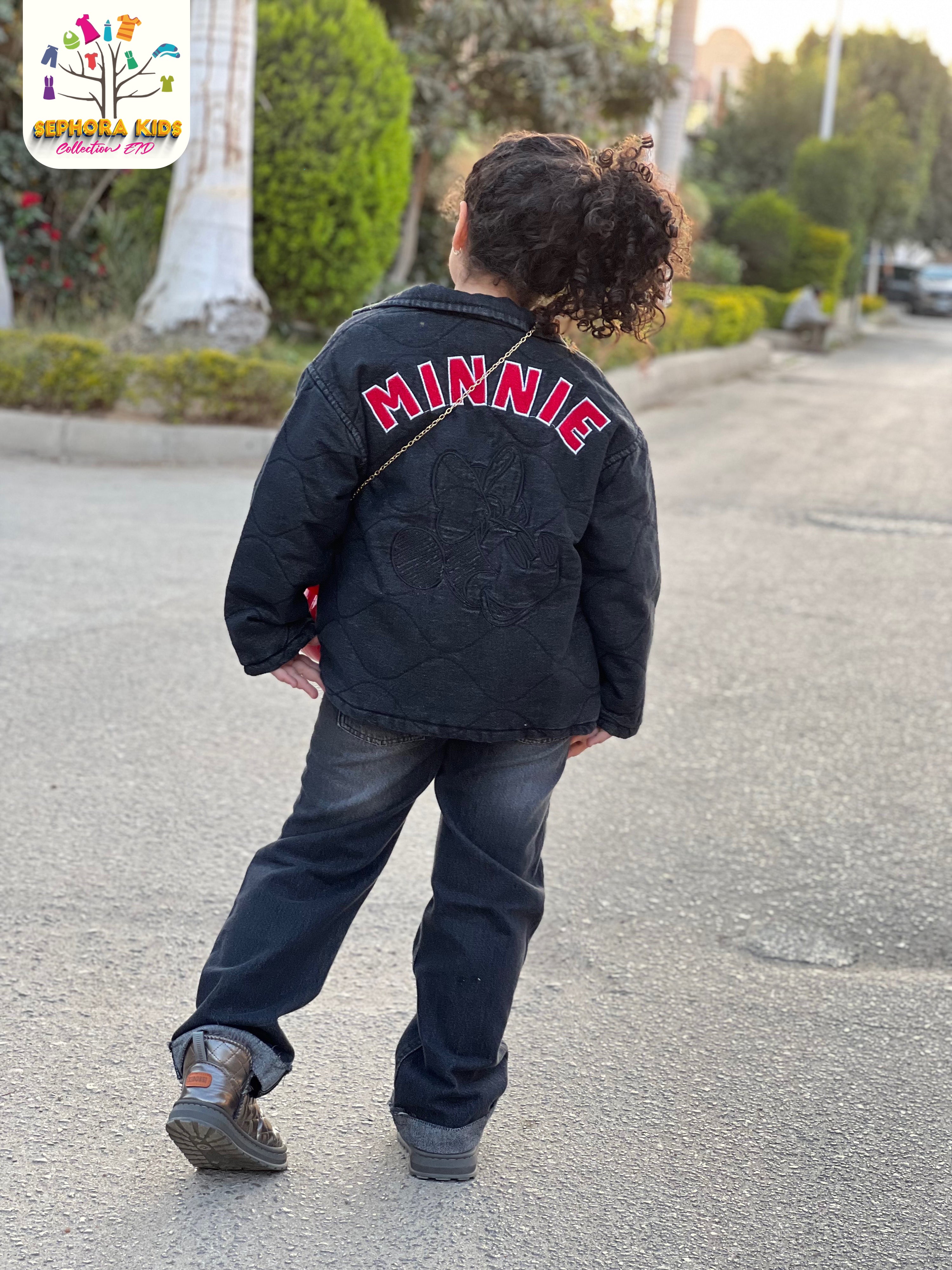 Child wearing a black jacket with 'MINNIE' on the back, walking on a street.
