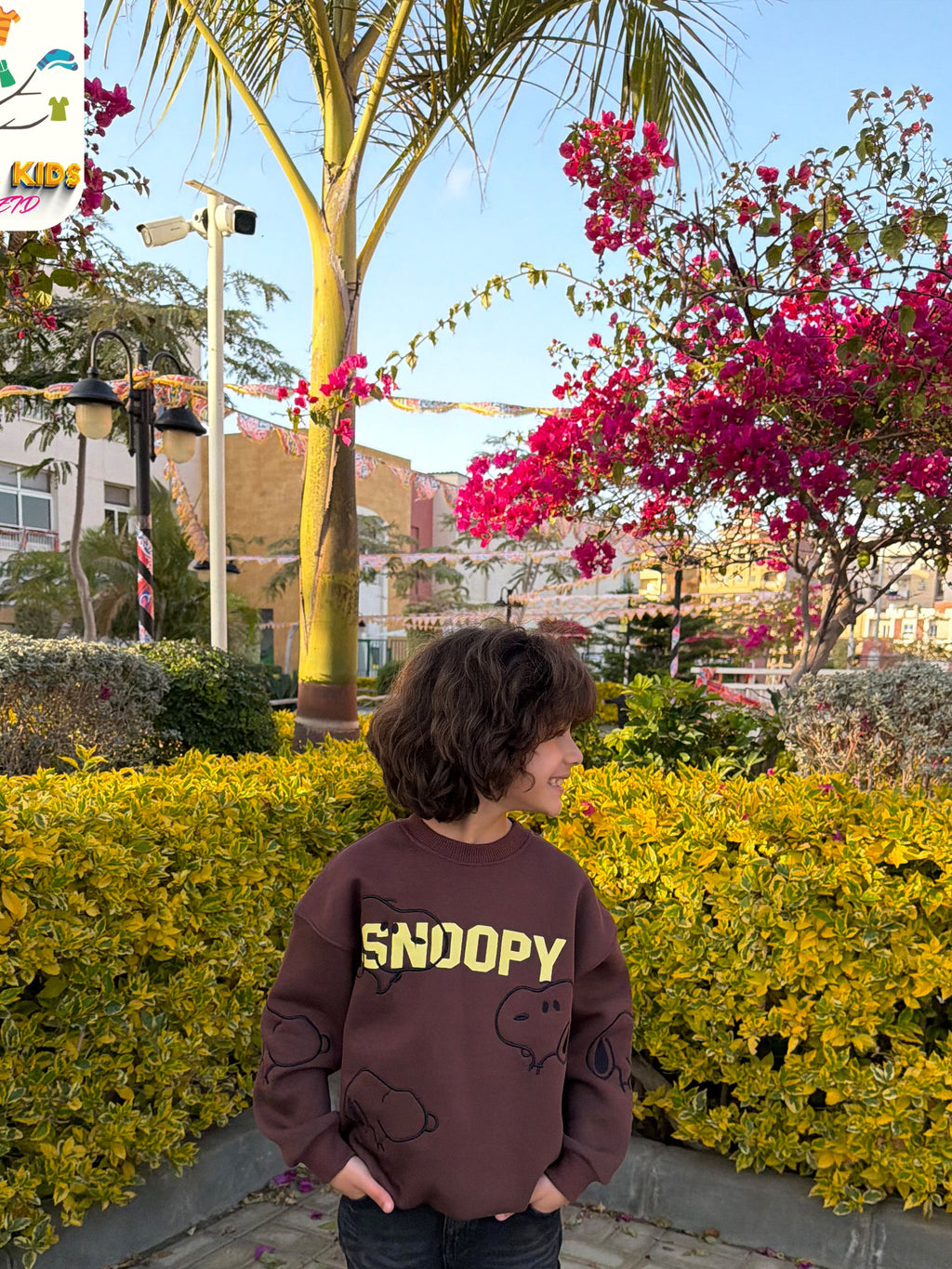 Child wearing a brown 'SNOOPY' sweatshirt standing in front of yellow bushes