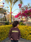 Child wearing a brown 'SNOOPY' sweatshirt standing in front of yellow bushes