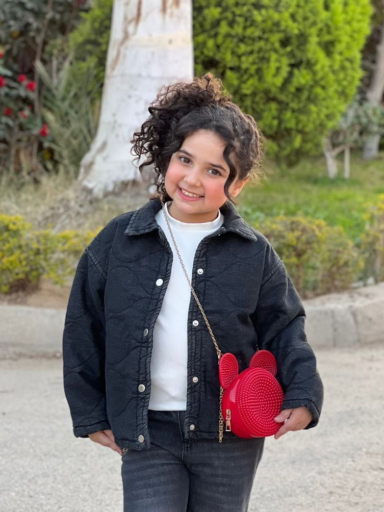 Child wearing a black jacket and jeans, holding a red heart-shaped bag outdoors.